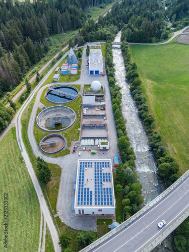 Aerial view of a wastewater treatment plant with circular sedimentation tanks, solar panels, and a flowing river in Welsberg-Taisten, Trentino-Alto Adige, Italy.