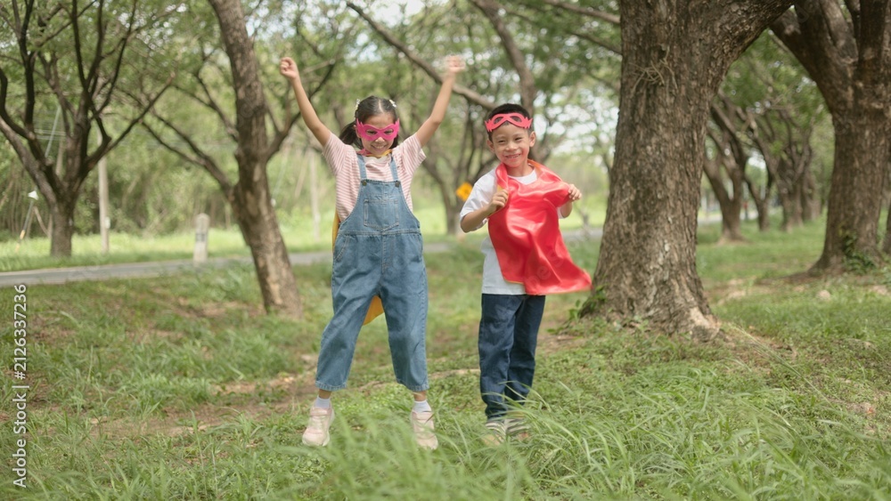 Fototapeta premium An excited Asian boy and girl jumping happily together while wearing colorful superhero capes during outdoor recreation time in a green park environment. area area area area