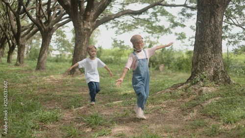 Canvas Print Two happy Asian children in superhero costumes running through the grass together while enjoying creative roleplay games in a public park setting