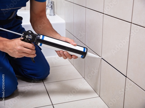 A construction worker in a blue uniform is applying silicone caulk to the tile wall in a bathroom.
