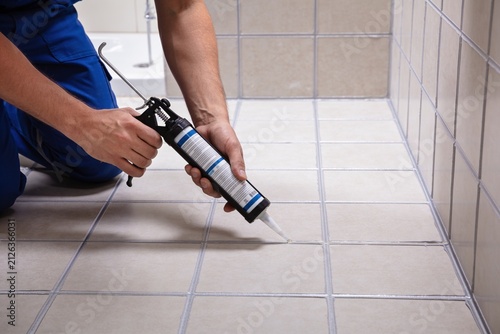 A close-up shot of a worker using a caulk gun to apply sealant between tiles in a tiled shower area.
