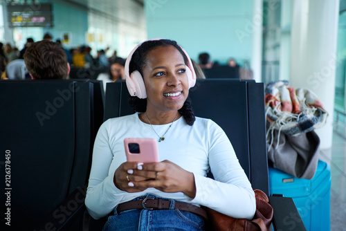 Young woman using phone to be connected and enjoying music on headphones, waiting for flight at a modern airport.