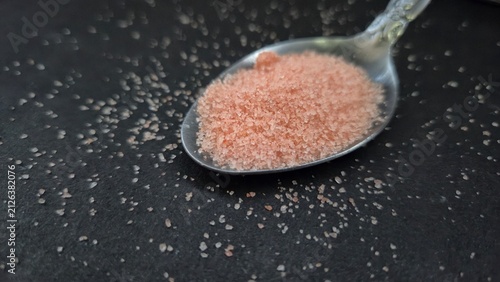 Close-up of fine pink Himalayan salt crystals in a silver spoon on a dark stone surface. The soft texture and natural mineral color create an elegant culinary and seasoning concept.