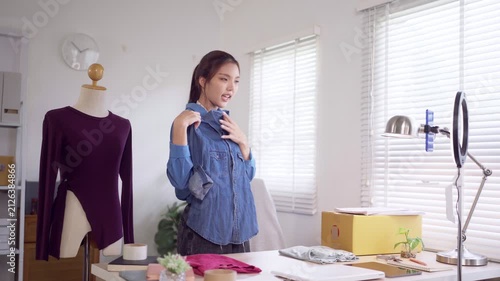 Young Asian online seller presenting blue denim shirt to camera while live streaming product from home studio