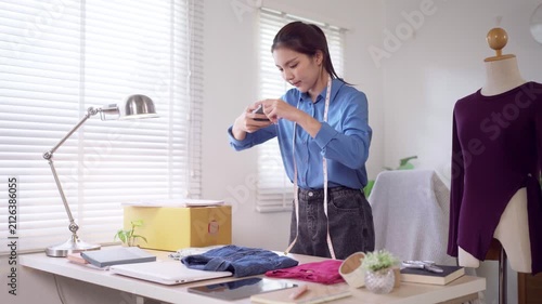 Young Asian fashion seller photographing clothing merchandise with smartphone while managing online boutique at home workspace