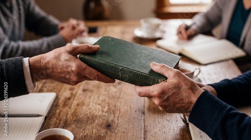 Sacred Exchange: An aged hand passes a worn, green sacred text to another, symbolizing the transfer of faith and wisdom during a solemn gathering.