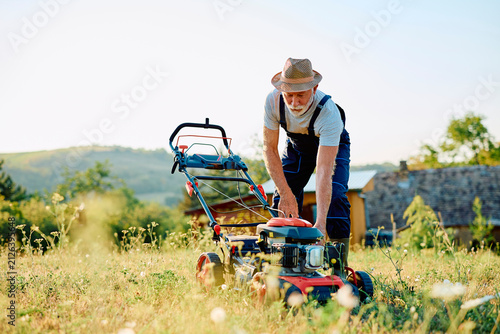 Foto Senior man in overalls and straw hat preparing a lawn mower for gardening work o
