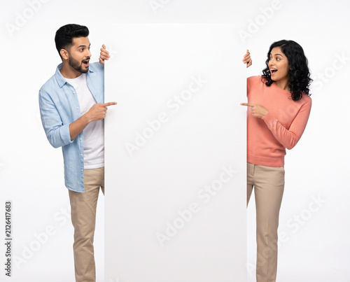 Young Indian couple peeking from opposite sides of a blank vertical display board while pointing toward the empty center with cheerful expressions isolated on a white background for promotional design