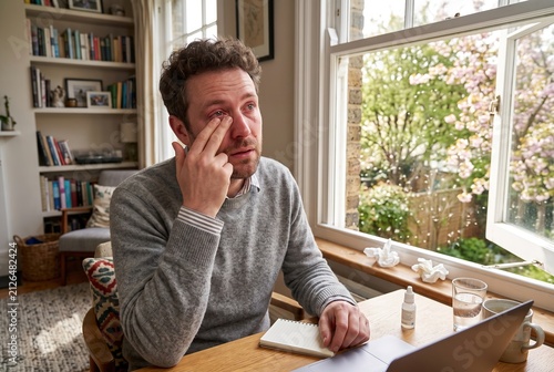 A man is sitting at a wooden desk by a large window in his home office. He is applying eye drops and seems to be deep in thought. Flowers can be seen outside