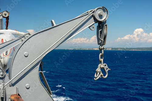 Foto Ship crane mechanism with a large metal hook and heavy chain hanging over the water, showing marine industrial equipment and the deep blue ocean waters landscapes