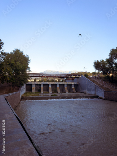 Urban Flood Control Dam with Pedestrian Bridge and Flowing River at SunsetA modern flood control dam with cascading water, a pedestrian bridge, and riverside infrastructure surrounded by lush trees un