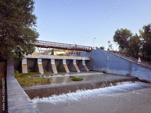 Fotografie Urban Flood Control Dam with Pedestrian Bridge and Flowing River at SunsetA mode