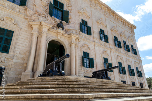 A baroque facade of Auberge de Castille rises above curved steps flanked by cannons, with a flag flying atop in Valletta, Malta