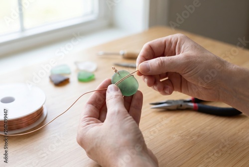 Hands wrapping green sea glass with copper wire to make jewelry on a wooden desk