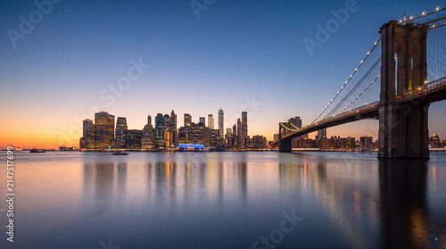 Panoramic sunset view of the New York City skyline with the Brooklyn Bridge illuminated over the East River during twilight hour with city reflections
