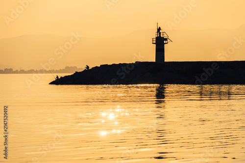 Lighthouse silhouette on a breakwater at golden sunset reflected on calm sea, coastal evening and travel destination.