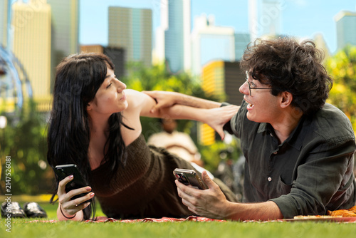 Fotografie Young man playfully teases his girlfriend and reaches toward her as they use smartphones and laugh on a picnic blanket, showing shared humor, affection, and a lively outdoor date in an urban park
