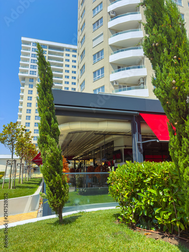 Canvas Print People enjoy their time at a restaurant near tall buildings with green trees and