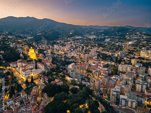 Aerial drone view of Sanremo cityscape and hills featuring the brightly illuminated Sanctuary of Madonna della Costa during twilight sunset in Liguria Italy