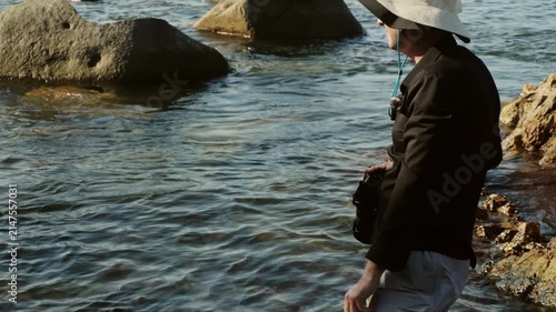 Young Caucasian male photographer is working outdoors on beach holding and using his camera with professional precision. He is in the middle of a fashion photoshoot at the beach capturing images