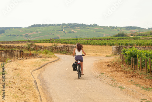 Woman cycling beside lush vineyard rows under a bright summer sky in Burgundy, France, surrounded by green grapevines, scenic hills, and rural countryside beauty.