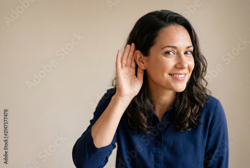 Smiling woman holds a hand to her ear to listen closely to a quiet sound or secret conversation