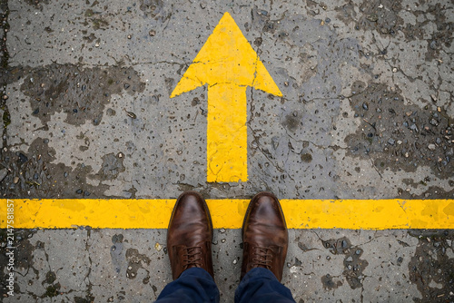 Brown leather shoes standing on wet asphalt with a bright yellow arrow pointing forward, top view, ideal for business strategy, forward movement and career progression concepts