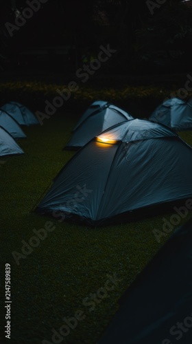 At dusk, the camping tents on the green grass are illuminated by local industrial outdoor lights, with a close-up shot of the warm light.
