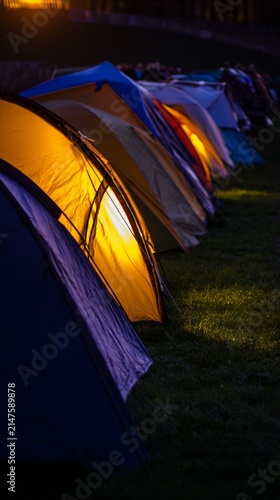 At dusk, the camping tents on the green grass are illuminated by local industrial outdoor lights, with a close-up shot of the warm light.