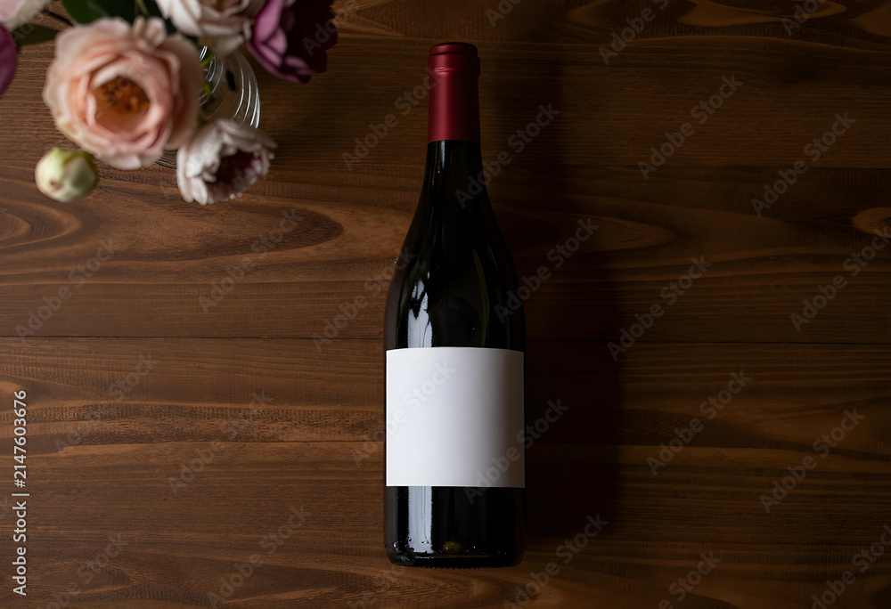 Obraz premium Red wine bottle with blank label and flowers on a dark wooden table, top view for branding and celebration concepts