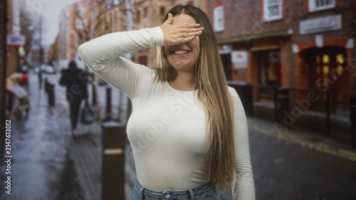 Woman covering eyes with hand and smiling on a wet urban street lined with brick buildings and pedestrians; playful shyness.