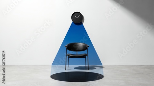 A sleek black chair sits beneath a dark wall clock, bathed in a conical blue light against a bright white studio wall.