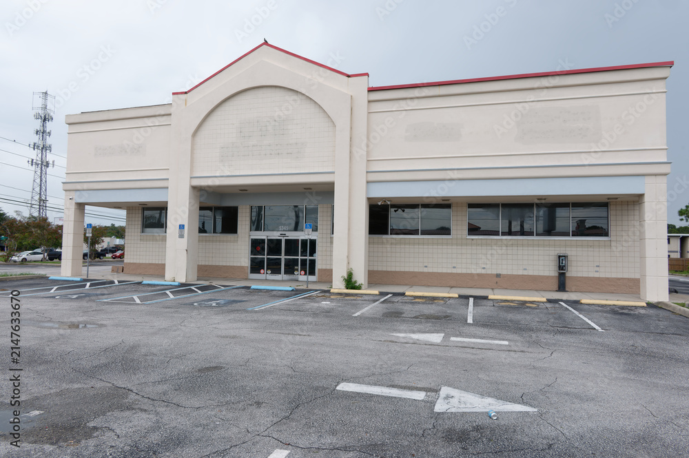 Fototapeta premium Empty vacant storefront in a closed retail building with an abandoned parking lot and overcast sky, conveying urban decay, economic decline and quiet commercial isolation in a suburban shopping center