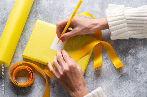 Person writing a card on a gift with yellow paper and ribbon on grey background.