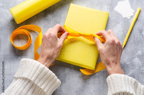 Hands tying a yellow ribbon on a yellow wrapped gift box on grey background.