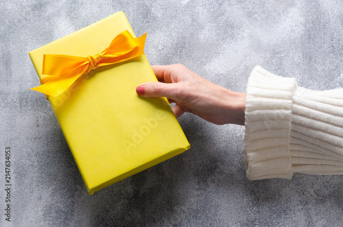 Woman's hand holding a yellow gift box with yellow ribbon on grey table with copy space. Horizontal format. 