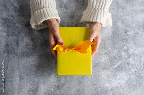 Woman's hands holding a yellow gift box with yellow ribbon on grey table with copy space.