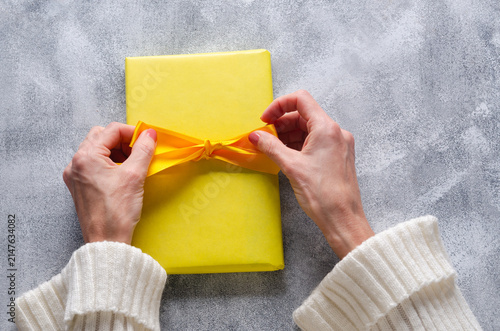 Person tying yellow ribbon on gift box on grey background. 