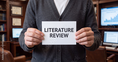Literature review sign held by professional man in business office setting wearing grey sweater demonstrating academic research and analysis concepts