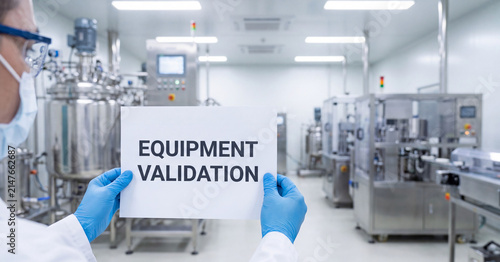 Equipment validation sign held by pharmaceutical worker wearing blue gloves and protective mask inside a sterile modern medical manufacturing laboratory facility