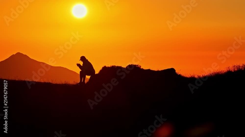 Solitary man sitting on a mountain summit using a smartphone against a beautiful orange sky in Col d'Aubisque in the French Pyrenees