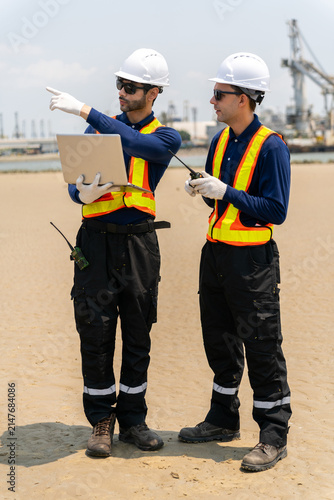 Two male engineers in safety vests and helmets discussing project details at shipyard port for logistics and industrial operations.