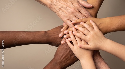 diverse hands of different ages and skin tones stacked together in a supportive gesture, soft studio lighting from above, neutral solid background with generous empty space around the hands