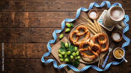 Oktoberfest flat lay background with fresh pretzels, beer mug, hops and blue ribbon on wooden table
