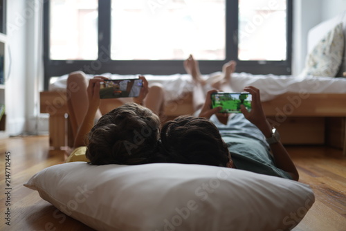 Two young boys lying on a wooden floor in a cozy bedroom, playing video games on their smartphones, sharing a moment of childhood friendship and screen time