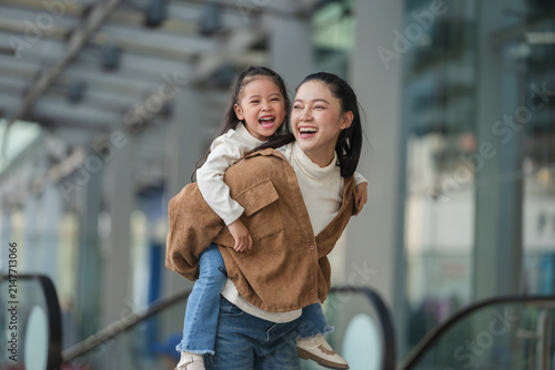 happy mother giving daughter piggyback ride near escalator in shopping mall or department store