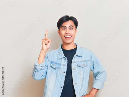 Excited young Asian man pointing his index finger up with a great idea while standing against a neutral background in a casual denim jacket.
