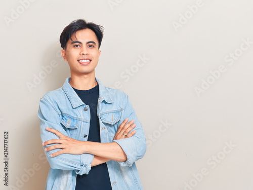 Confident young Asian man standing with arms crossed and smiling at the camera in a studio while wearing a denim jacket for a casual lifestyle portrait.