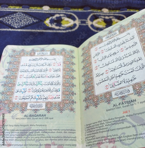 Canvas Print An open Quran showing Surah Al-Fatihah and Al-Baqarah rests on a navy blue prayer mat with maroon-gold decorative borders, capturing a quiet moment of reflection
