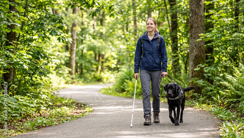 Visually impaired woman with guide dog and white cane walking on a forest path.